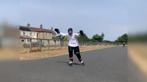 Andy Smart Will skating with his white skate top, black trousers and knee guards and inline skates on. He has his hands, with gloves on, in the air and is wearing his helmet. He is skating along a paved area. 
