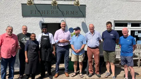 A line of nine people standing in front of the Fishers Arms, which is a white pebble dash building with a grey sign above the door. David Smith MP is standing in the centre and has grey hair and is wearing a pink shirt and dark trousers and brown shoes with his hands clasped at his front. To the left is William and Mary Ndeme in black aprons, William is wearing a black hat. The others are all men who are smiling into the camera.