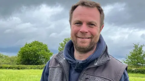 Olly, wearing a blue hoodie and grey gilet, is standing in the middle of a field on a cloudy day. 