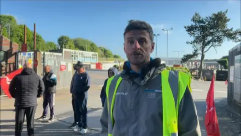 A man wearing a grey jacket and reflective vest stands at the Lifford Lane depot. A group of men can be seen standing in a line behind him.
