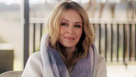 Kylie Minogue smiling at the camera while wearing a fur coat in the new three-part Netflix documentary, Kylie
