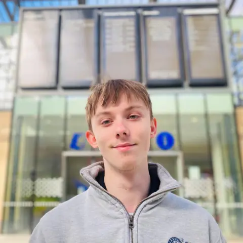 Man - Luke Iden - smiling at camera, wearing a grey zipped top, inside train station.