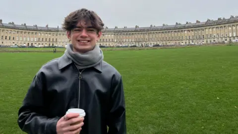 Fern Sherwood Sam, smiling into the camera and holding a takeaway coffee. His black leather jacket is zipped up and he is standing on the grass in front of the iconic Royal Crescent in Bath.