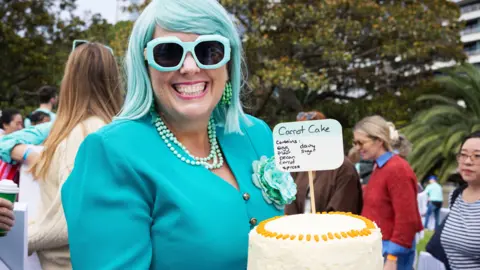 A woman with turquoise hair and glasses, green earrings and necklace and a bright turquoise top, with green and turquoise bangles, holding a carrot cake