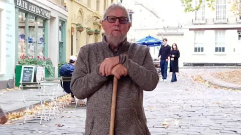 A man stands in the centre of a cobbled street. He is wearing a thick brown jumper and is leaning on a brown stick. He has grey hair and glasses and behind him are a man and woman walking. There is also a shop with a sign and tables and chairs. 