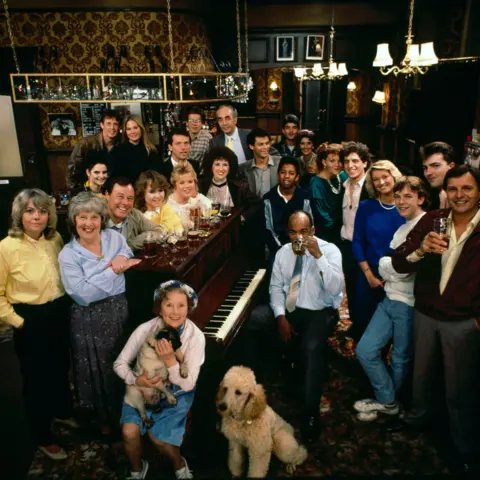 BBC The cast of EastEnders in 1985, sat around the Queen Vic piano