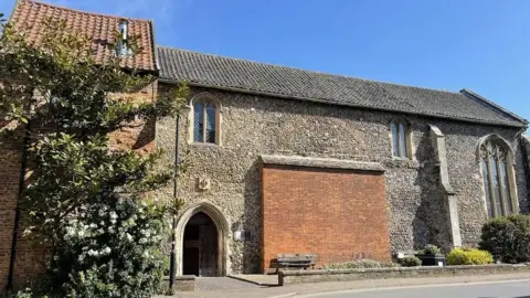 The outside of Becket's Chapel in Wymondham. It is a large stone building with arched windows and a tiled roof. There is a large shrub growing up one side.