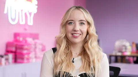 A woman with long blonde hair in a black and beige shop, with a pink-purple blurred background