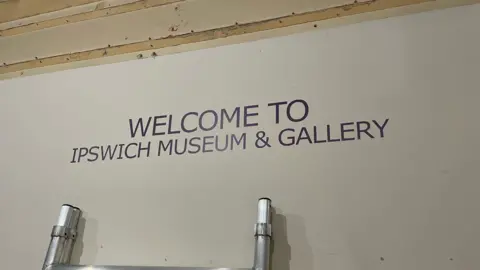 Shaun Whitmore/BBC A sign is pictured which reads, 'Welcome to Ipswich Museum & Gallery'. The writing is in a black font on a white background. The top of a ladder can be seen resting underneath.