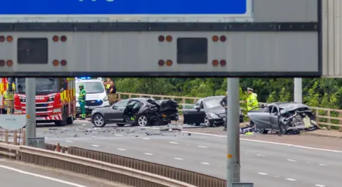 Robert Perry A crash scene on a motorway, with three cars smashed up. Police and other emergency services members are standing around the scene, with bits of the cars lying on the road.