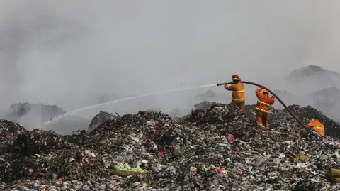 Getty Images Firefighters in orange uniforms standing on a gigantic pile of rubbish, holding a hose and spraying water, as smoke billows into the air