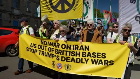 Protesters at the front of a demonstration are holding a large yellow anti-war banner, with many people following behind them.