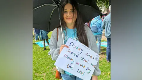 Lauren A teenage girl with dark hair stands on grass. She is wearing a blue t-shirt with Widows Fight on the front, a grey zip up jumper and blue jeans. She is holding an open black umbrella above her head and a sign saying "Daddy died scared about how Mummy would pay the bills"