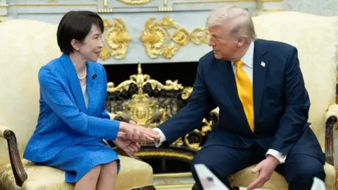 US President Donald Trump shakes hands with Japanese Prime Minister in the Oval Office. Both are seated and wearing blue suits