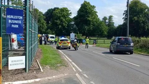 A number of police and ambulance vehicles are at the scene of a crash. In the foreground is the entrance to a business park. A police motorbike stands next to a police car in the centre, on the road