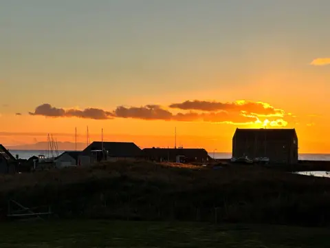 William Hardie A harbour at sunset with the building silhouetted by an low orange winter sunset and the masts of a few boats sticking up too