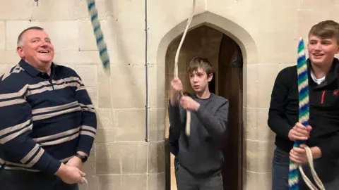 Three people pulling on ropes that lead up to a ceiling. They are standing in an indoor church setting.