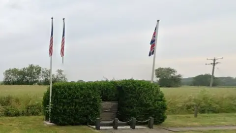 Google Small inscribed stone surrounded by hedge. There are three flags around the memorial on the edge of an airfield.