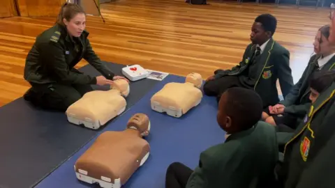 Ross Miklaszewicz/BBC Students sit on the floor as they are taught by a LAS medic