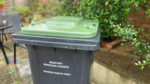 Bedford Borough Council A black bin with a green lid is sitting on a patio in front of a low wall and some shrubs. On the bin is printed "Bedford Borough Council garden waste only" in white, capital letters