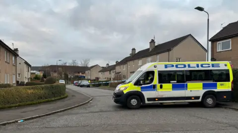 PA Media A police van blocks off a residential road. Police tape can be seen stretching across the road. The houses are covered in grey render. 