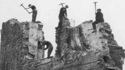 A black and white image of a demolition site of workers breaking down the old prison site. Lots of rubble and walls left.