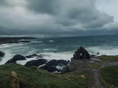 Scott Innes Moody landscape image of ruins of a building next to dark grey waters as a storm comes in