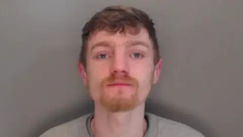 Mugshot of Rhys Smith. He has short messy dark hair and a thin ginger goatee and is wearing a grey sweatshirt.