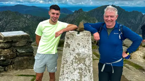 Robert Marshall Ross is wearing a yellow T-shirt and grey shorts. Robert is in a blue top and dark trousers. They are smiling. Robert has white hair, Ross has brown hair. They are standing beside a cairn on top of a mountain. There are the tops of mountains in the background.