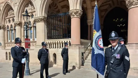 Jenny Kirk/BBC A police guard of honour stand in two lines facing each other outside a large civic building on a sunny day. One officer stands with a large blue flag with a police emblem on it and looks at the camera. All the officers are wearing matching uniform with black hats, black suits and trousers and white gloves. Some stand with their hands behind their backs while another to the left of the image has his hands in front of him as he holds a brown staff.