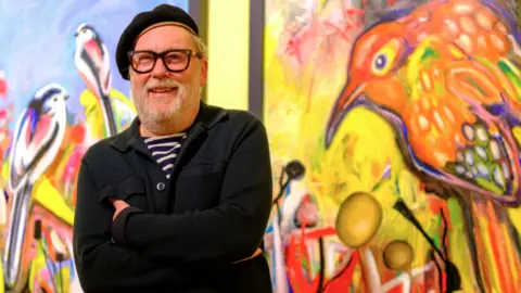 Jim Moir smiles at the camera, with two of his vivid paintings of birds hanging behind him. He is wearing a black beret, striped t-shirt, dark overshirt and glasses.