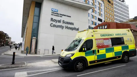 An ambulance drives past the Royal Sussex Hospital in Brighton. It is a modern building with the name of the hospital on the side in large letters.