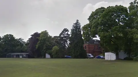 Google A cricket pitch bordered by large trees, with Leahoe House and parked vehicles partly visible in the background. A single-storey brick pavilion is visible to the left, alongside a sight screen. A large country house is visible behind trees.