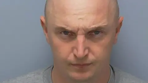 Adam Wright frowns at the camera in a police custody photo. His head is shaved and he wears a grey, collarless top.