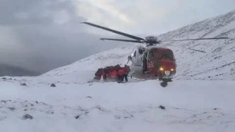 Members of Lochaber MRT bring a stretcher to a coastguard helicopter during a rescue in March. The team, dressed in red jackets, crouch down due to the down draft from the helicopter's rotors. The rescue is taking place in a snowy, mountain landscape.