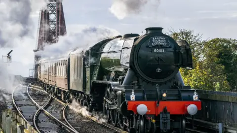 The Flying Scotsman train. It is a green steam train with a plaque on the front reading "Railway Touring Company"