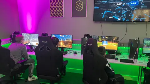 Three boys sit on black gaming chairs in front of computer monitors playing different games. The room is lit with pink and green lights. There is a large screen on the wall and two posters.