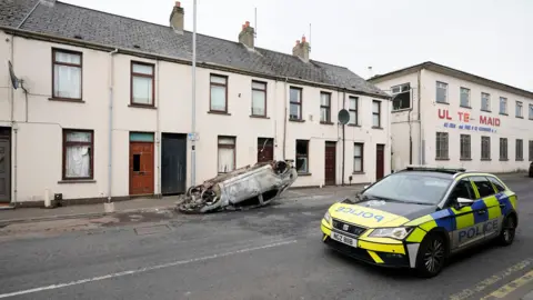 PA Media An overturned and burned car sitting in front of a row of houses in Ballymena. A police patrol car is parked opposite.