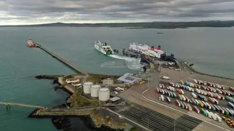 A concrete dock, with two boats in the sea in front of it, and a car park to one side, full of vehicles, with various industrial buildings on the other side of the dock
