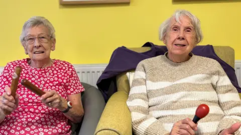 An older woman with grey hair and glasses, wearing a pink flowery top, and a grey-haired woman in a brown and white striped jumper sitting o armchairs with a yellow wall behind