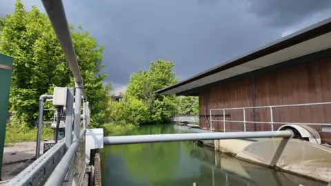 Osney Lock Hydro has a body of water running through, with mechanical equipment on one side and a wooden building on the other.