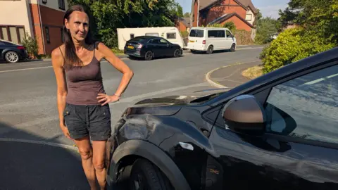 Zoe Farmer Zoe Farmer stood beside her black car which has scratches and dents along the front. She has brown hair and is wearing a brown top with black shorts.