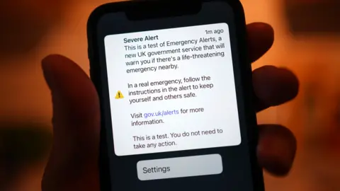 A mobile phone screen held in someone's hand displays a test emergency alert message.