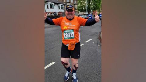 Paul is running in an orange race shirt with his race number pinned to the front. He has both thumbs up and is smiling. You can see his stoma bag also with an orange cover.