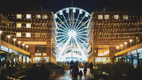 Coventry City Council A giant observation wheel in a city centre, with Christmas lights on the buildings and a church tower in the far background 