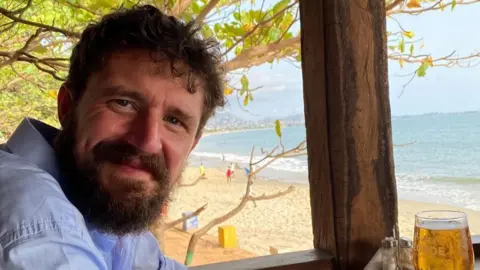 Jersey Overseas Aid A smiling Simon Boas sitting in a beach hut with a drink in front of him on a table