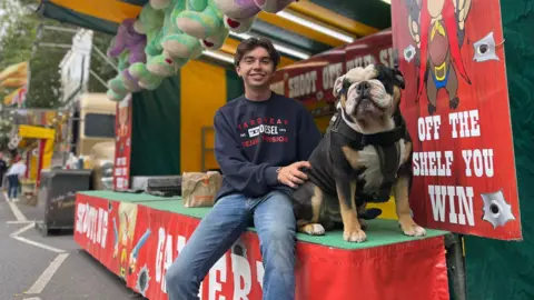 A young man sits with his dog in front of a Western-themed shooting range at the fair.