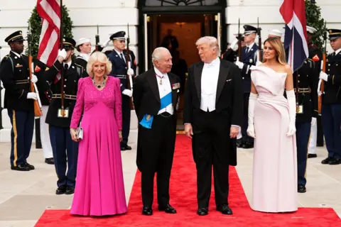 PA Media The two couples stand outside the entrance to the White House with soldiers and guards and flags around them standing on a red carpet.