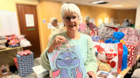 BBC Helen Wood stands holding a blue gift bag on which can be seen a smiling bunny, while in the background an array of presents are stretched out across the room 