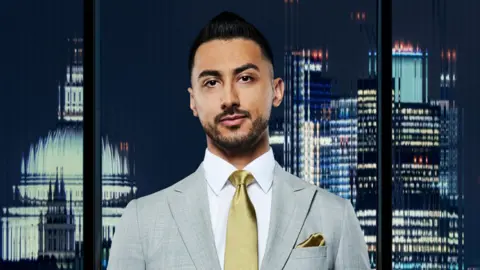 Rajan Gill in a grey suit with a gold tie. A dar kcity skyline is seen in the background.